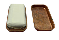 A vintage rectangular metal tray with a warm copper or bronze-toned interior and rim, shown open with the lid removed. One side contains a folded off-white paper wrapper or cover, while the other side shows the textured, aged metal base with embossed text or markings.