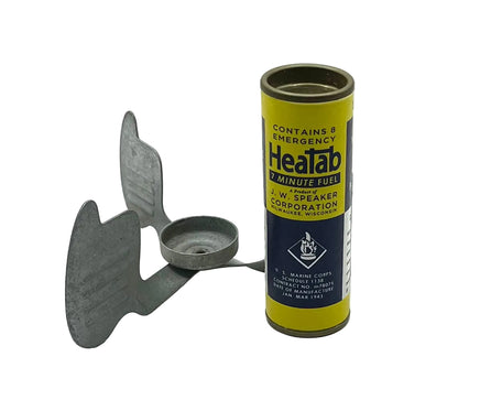 A metal emergency stove with a three-blade wing design stands next to its yellow and navy blue cylindrical canister labeled 'Heatab 7 Minute Fuel' on a white background.