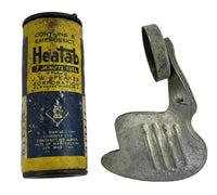 A vintage Heatab emergency fuel canister next to a metal clip or hook, shown on a plain light background. The canister is yellow and blue with black text, and the metal clip is silver.