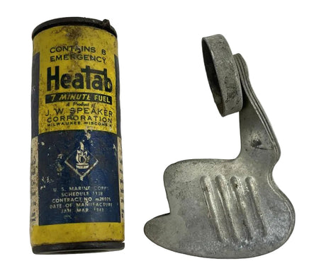 A vintage Heatab emergency fuel canister next to a metal clip or hook, shown on a plain light background. The canister is yellow and blue with black text, and the metal clip is silver.